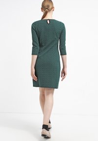 Vihreä neulemekko, jossa on kolme neljäsosaa hihat ja takana avaus. Kangas on tekstuurilla ja muoto on vartalonmyötäinen.