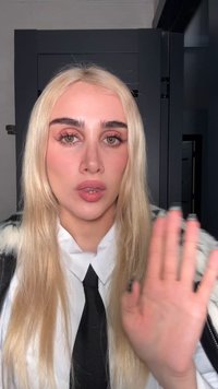 Portret osoby z długimi, prostymi blond włosami, ubraną w białą koszulę z kołnierzykiem i czarnym krawatem, z podniesioną ręką wykonującą gest.