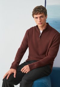 Bordový sveter na zips s rebrovaným úpletom, s vysokým golierom a dlhými rukávmi, skombinovaný s tmavými nohavicami, sediaci na modrom povrchu.
