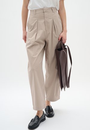 Person wearing beige pleated trousers, black loafers, white top, holding a dark brown leather tote bag by the handle.