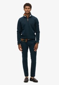 Marineblaues Fleece-Pullover mit Reißverschlusskragen, über ein kariertes Hemd getragen. Slim-Fit dunkle Hose und braune Slipper runden das Outfit ab.