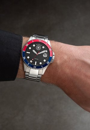 Montre-bracelet avec lunette rouge et bleue, cadran noir, index argentés, date affichant le 22, et bracelet en acier inoxydable au poignet gauche.