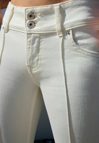 Bijele traperice visokog struka imaju dva srebrna dugmeta na pojasu, glatku teksturu i klasičan dizajn s pet džepova, uz suptilno šivanje.