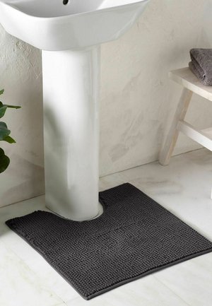 Tapis de bain texturé gris foncé avec une découpe ajustée autour d'un lavabo sur pied blanc sur un sol de salle de bain carrelé clair.