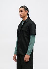 Černá krátká košile s krátkým rukávem a dvěma předními kapsami, nošená přes tyrkysový top s dlouhým rukávem. Košile má zapínání na knoflíky a volný střih.