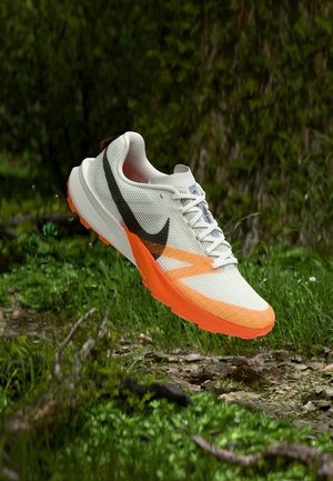 Chaussure de course blanche et orange avec le logo Nike noir, en suspension au-dessus d'un sentier rocheux et herbeux dans un cadre forestier.