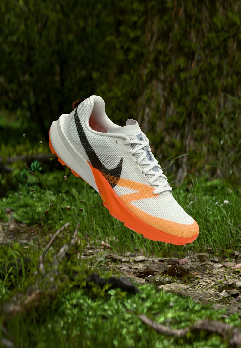 Λευκό και πορτοκαλί αθλητικό παπούτσι με μαύρο λογότυπο Nike, στον αέρα πάνω από ένα βραχώδες, γρασίδι μονοπάτι σε δασικό περιβάλλον.