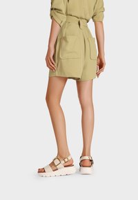 Completo beige a due pezzi composto da una camicia con cintura e pantaloni corti fino al ginocchio con grandi tasche posteriori. Sandali bianchi spessi con accenti dorati.