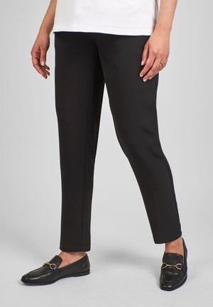 Kvinde iført sorte skræddersyede bukser, sorte loafers med gulddetaljer og en hvid top, stående mod en ensartet lys baggrund.