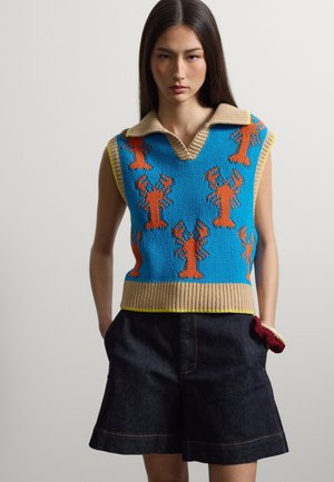Jeune femme portant un gilet bleu tricoté sans manches avec des homards orange et un col beige, associé à un short en denim foncé.