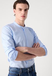 Šviesiai mėlynas, ilgomis rankovėmis marškinis su juostelėmis ir mygtukais; lygus paviršius, prigludęs dizainas. Modelis pozuoja su rankomis sukryžiuotomis, vilkėdamas džinsus.