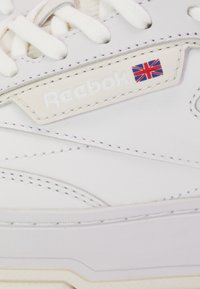 Beli usnjeni športni copat z teksturirano površino, Reebok logotipom in detajlom britanske zastave. Šnure in šivi v usklajeni beli barvi.