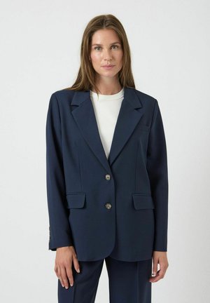 Blazer entallado azul marino con dos botones, solapas con muesca, dos bolsillos frontales y un tejido sutilmente texturizado. Se lleva sobre una blusa blanca.