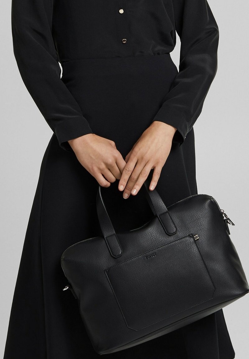 Personne vêtue d'une tenue noire tenant un sac à main en cuir noir avec doubles poignées et une poche avant sur fond gris.
