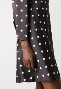Abito in satin nero con pois bianchi; presenta maniche lunghe e dettagli con bottoni vicino al polsino; texture morbida.