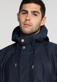 Navy waterproof jacket with large hood, silver snap buttons, drawstring, and smooth texture. Features a minimalistic design with contrasting inner lining.