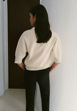 Femme aux longs cheveux noirs portant un pull crème à manches courtes et un pantalon noir, debout avec les mains derrière le dos contre un mur blanc.