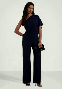 Mulher vestindo um macacão azul marinho de um só ombro, sapatos de salto abertos pretos, segurando uma carteira preta, de pé contra um fundo simples.