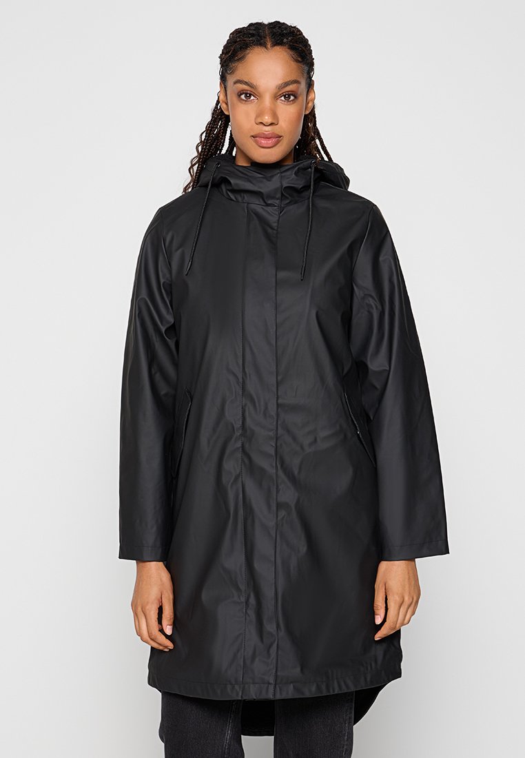 Femme portant un long imperméable noir à capuche avec des poches à l'avant, se tenant devant un fond clair.