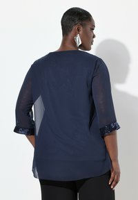Marineblaue Bluse mit Dreiviertelärmeln, transparentem Stoff und mit Pailletten verzierten Bündchen. Weiche Textur, lockere Passform und abgerundeter Saum.