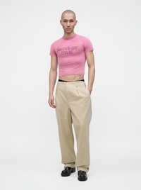 Άτομο φοράει ροζ crop top με κείμενο, μπεζ παντελόνια με φαρδιά πόδια και μαύρα παπούτσια, στέκεται μπροστά σε απλό φωτεινό φόντο.