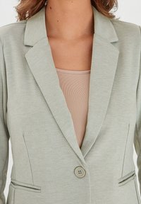 Blazer verde chiaro con rever, chiusura a singolo bottone e tasche laterali. Realizzato in tessuto testurizzato, sovrapposto a una maglietta beige.
