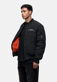 Giubbotto bomber nero con fodera arancione, tasca con zip sulla manica, polsini a costine e testo ricamato sul petto. Indossato da un modello.