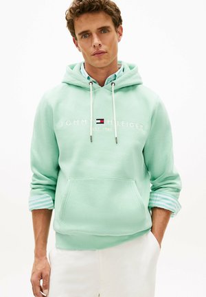 Hombre joven con sudadera con capucha Tommy Hilfiger de color verde menta con cordones blancos y pantalones blancos, de pie frente a un fondo liso.