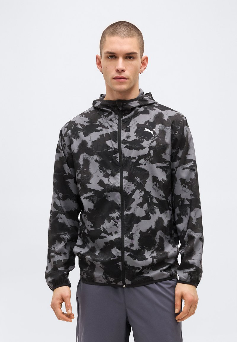 Jeune homme aux cheveux courts portant un sweat à capuche zippé camouflage noir et gris et un pantalon gris, debout devant un fond clair uni.