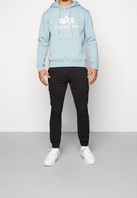Felpa con cappuccio azzurro chiaro con logo bianco, abbinata a pantaloni cargo neri con tasche laterali e sneaker bianche. Texture liscia su tutta la superficie.