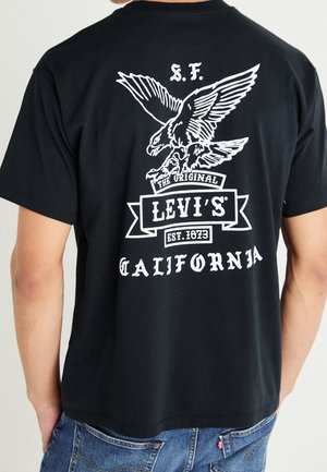 Person iført sort Levi's T-shirt med hvid ørn-grafik og teksten "S.F. The Original Levi's Est. 1873 California" på ryggen.