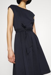 Námořnické modré šaty s krátkými rukávy, texturovaná látka, přiléhavý korzet a detail s uvázaným pasem. Rozšířená sukně sahá pod kolena.