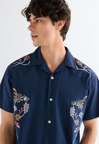 Νεαρός άνδρας με σγουρά σκούρα μαλλιά, φορώντας μπλε σκούρο κοντομάνικο πουκάμισο με πολύχρωμη κεντητή λουλουδάτη διακόσμηση στους ώμους και το στήθος, κοιτάζοντας προς τα αριστερά.