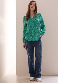 Blouse verte avec un motif texturé, décolleté en V et manches longues, associée à un jean bleu et des baskets blanches. Look simple et décontracté.
