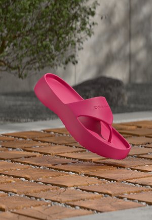 Șlăpri cu platformă roz aprins Calvin Klein, suspendați deasupra unui bloc de lemn și a unei suprafețe acoperite cu pietricele.