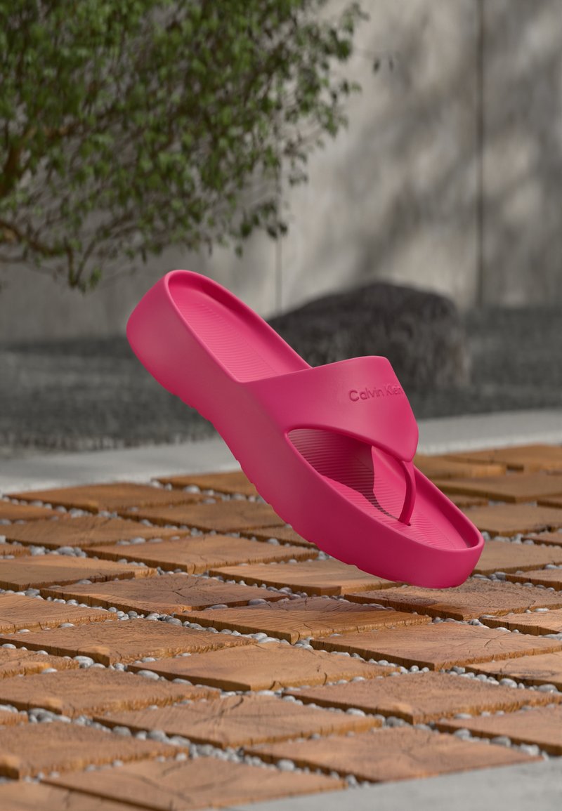 Φωτεινά ροζ πλατφόρμες σαγιονάρες Calvin Klein, κρεμασμένες πάνω από ξύλινο κούτσουρο και επιφάνεια με βότσαλα.