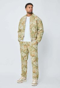 Homme se tenant debout les mains dans les poches, portant une veste et un pantalon à motifs floraux beiges et verts avec des baskets blanches et un t-shirt blanc.