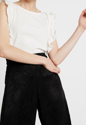 Valkoinen paita, jossa on röyhelösateen yksityiskohta, paritettu korkeavyötäröisten mustien housujen kanssa, joissa on aaltoileva tekstuuri. Sujuva kangas, rento istuvuus.