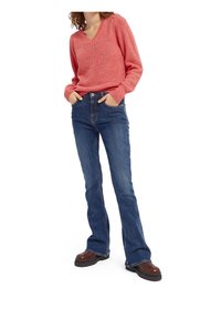 Suéter de punto texturizado rojo con escote en V, combinado con jeans de mezclilla flare azules y botas de suela gruesa marrones. Manos en los bolsillos.