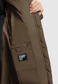 Brūns quiltēts jaka iekšpusē ar sānu kabatu, ar melnu uzlīmi baltā tekstā. Gluda, viegla auduma.