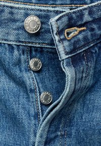 Close-up van denim jeans met een donkerblauwe stof, zilveren metalen knopen en een contrasterend gouden stiksel.
