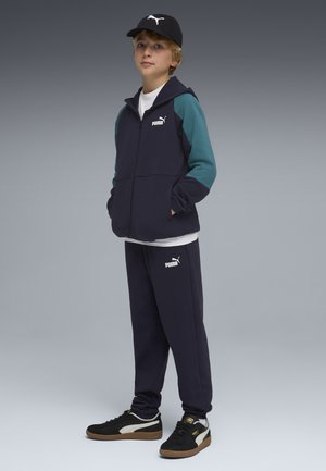 Ragazzo con tuta Puma blu navy e verde teal, berretta Puma nera e sneakers Puma nere, in piedi con le mani nelle tasche su sfondo grigio.