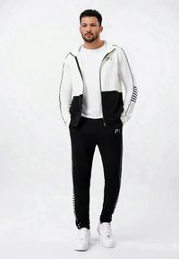 Tuta sportiva bianca e nera con giacca zip e pantaloni, caratterizzata da un logo e righe laterali, abbinata a sneaker bianche.