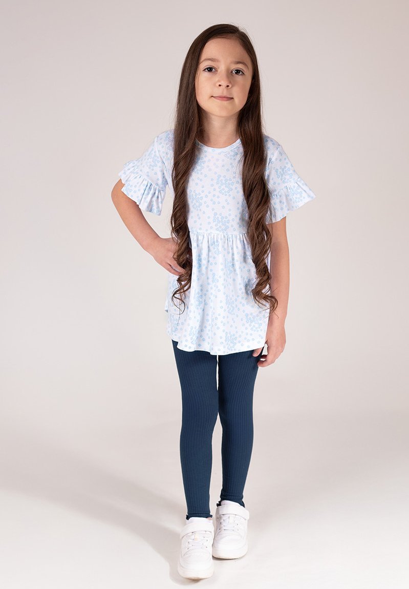 Jasnoniebieska sukienka z krótkim rękawem, falbanami na rękawach i talią z falbaną, zestawiona z granatowymi prążkowanymi legginsami i białymi trampkami.