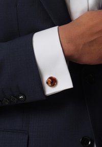 Manschettenknopf in runder Form mit einem braun-orange marmorierten Stein, der in einen polierten Silberrahmen eingefasst ist, angebracht an der Manschette eines weißen Hemdes.