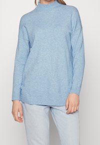 Pull tricoté bleu clair avec un col montant côtelé, des épaules tombantes et des poignets côtelés ; texture douce, coupe ample et longueur jusqu'aux hanches.