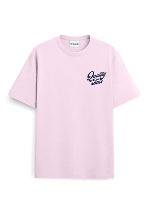Joven con pelo rubio rizado que lleva una camiseta rosa claro con el texto "Quality Time" y pantalones negros, de pie con las manos en los bolsillos.