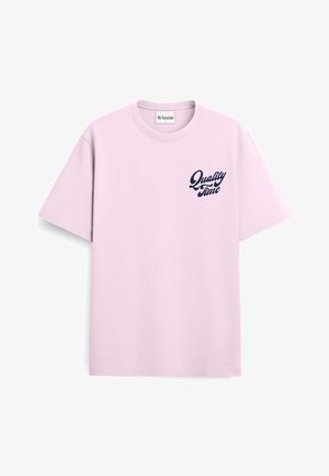 Młody mężczyzna z kręconymi blond włosami, ubrany w jasnoróżowy T-shirt z napisem „Quality Time” i czarne spodnie, stojący z rękami w kieszeniach.