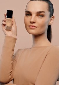 Base de maquillaje KIKO Unlimited en un frasco de vidrio esbelto con tapa negra. El modelo lleva una camiseta de manga larga beige ajustada, con un fondo neutro.