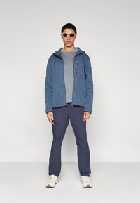 Μπλε υφασμάτινη ζακέτα με φερμουάρ και κουκούλα, γκρι μπλουζάκι, ναυτικό μπλε παντελόνι και ανοιχτόχρωμα παπούτσια. Το μοντέλο φορά γυαλιά ηλίου και στέκεται μπροστά σε μονόχρωμο φόντο.